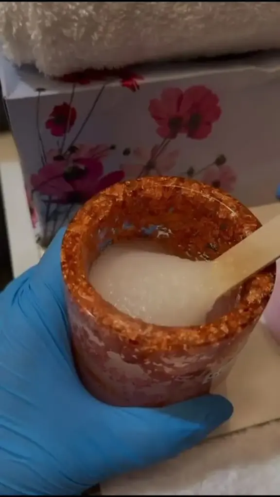 A jar of body scrub with a wooden spatula, held in a gloved hand