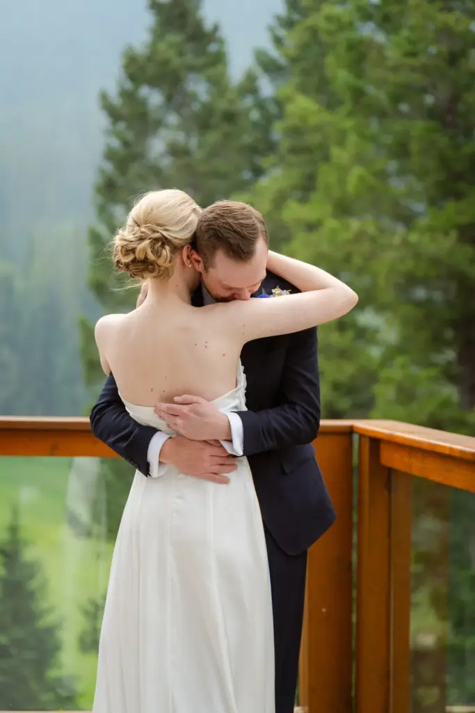 A bride with a blonde updo embracing the groom on a mountain deck
